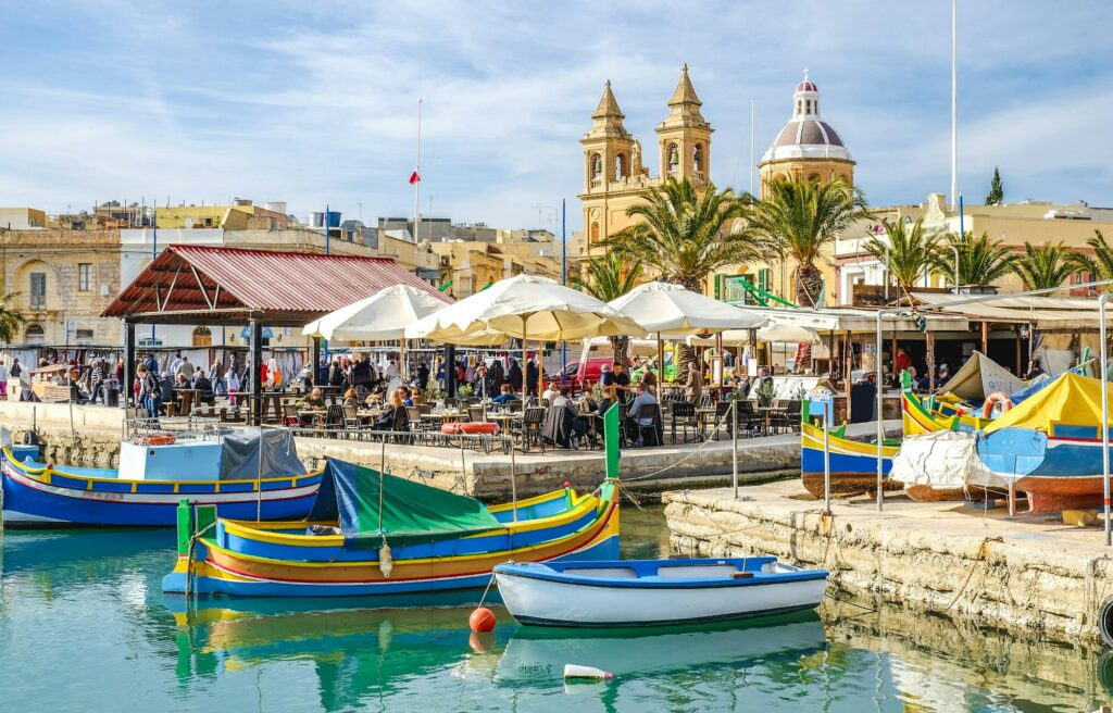 The Marsaxlokk Fish Market in Malta: Colorful luzzu boats bob in the harbor as fishermen unload fresh seafood onto bustling stalls. Visitors browse the catch of the day, enjoying the vibrant atmosphere of this iconic Mediterranean market.