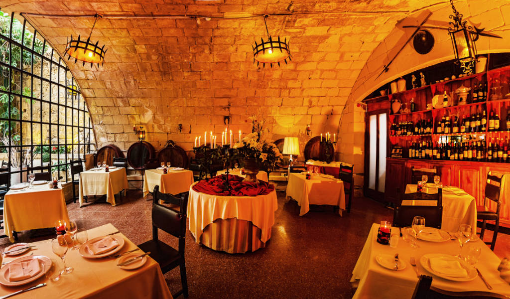 A charming courtyard adorned with lush greenery and vibrant flowers, serving as the outdoor seating area for Bacchus, a hidden gem restaurant in Mdina, Malta. Tables draped with crisp linens are surrounded by wrought-iron chairs, creating an inviting atmosphere for diners to enjoy their meals al fresco. Soft overhead lights illuminate the space, casting a warm glow as guests indulge in delicious cuisine and fine wines under the Mediterranean sky.