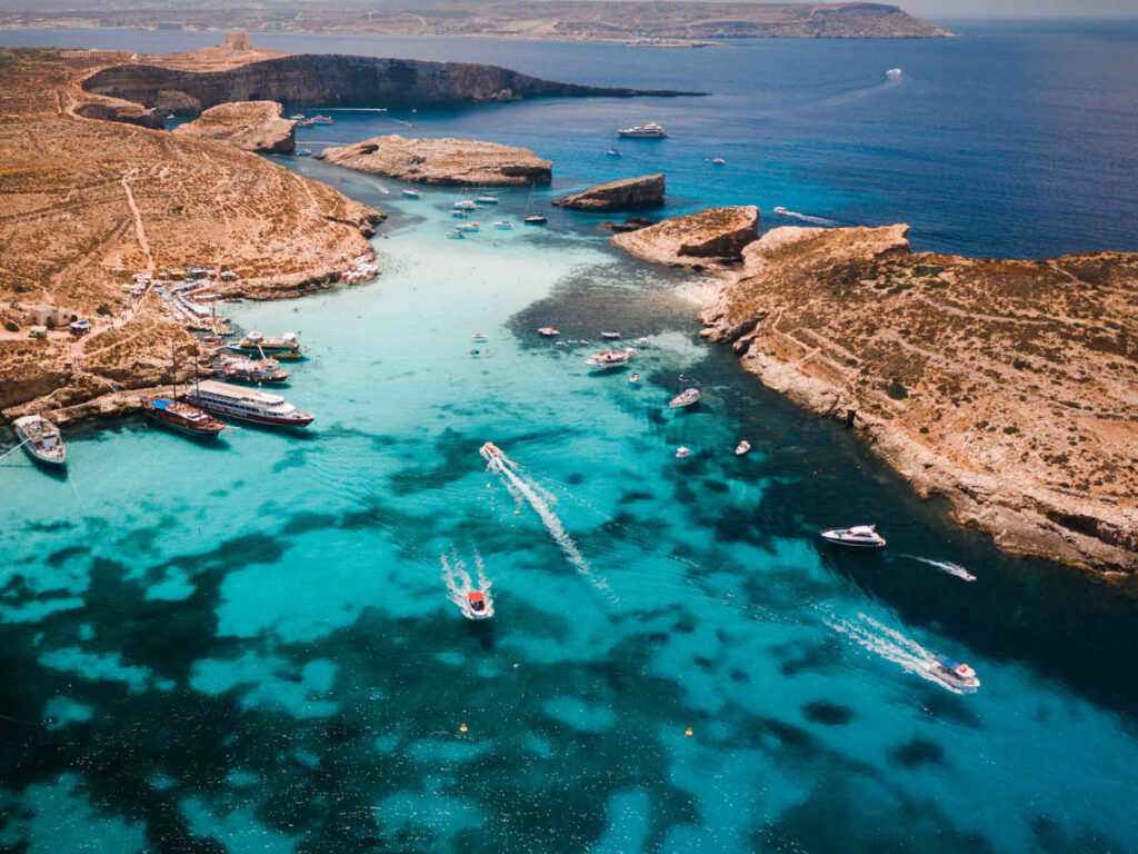 Aerial view of Comino Island, Malta: crystal-clear turquoise waters, rocky coastline, and sandy beaches. The uninhabited island is surrounded by the Mediterranean Sea, offering a pristine natural environment for swimming, snorkeling, and sunbathing.









