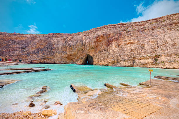 Dwejra, Gozo, Malta: A scenic view of the rugged coastline and limestone cliffs surrounding the picturesque inlet. The azure waters of the Mediterranean contrast with the golden hues of the cliffs, creating a breathtaking backdrop for sunset picnics and romantic strolls along the shoreline.