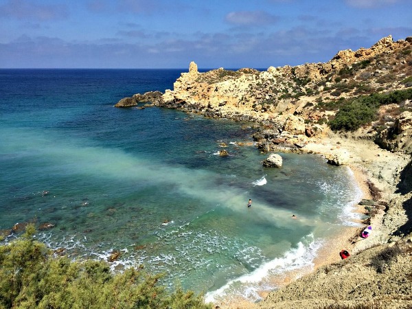 Fomm ir-Riħ, Malta: A secluded beach nestled between rocky cliffs, with clear blue waters lapping against the shore under a sunny sky.