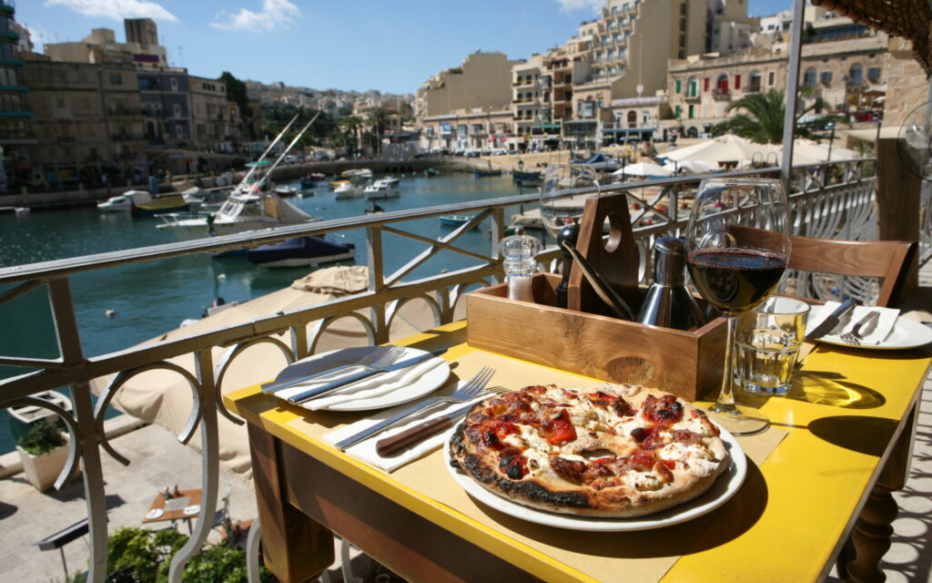 A picturesque scene at Gululu, with a mouth-watering pizza accompanied by a glass of wine, set against the backdrop of the sparkling sea, promising a perfect dining experience blending delicious cuisine with breathtaking views.