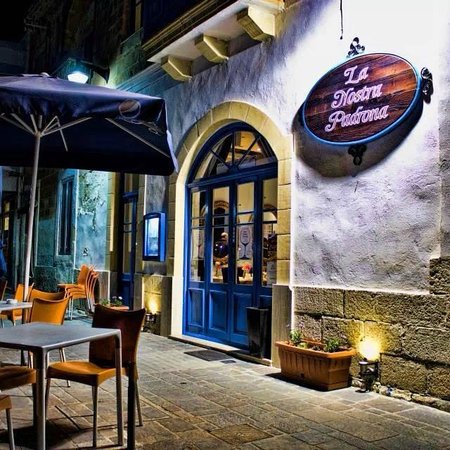 A cozy Mediterranean restaurant named La Nostra Padrona in Mellieħa, Malta. The restaurant's warm ambiance invites diners to enjoy delicious seafood dishes in a relaxed setting. Outdoor tables are adorned with crisp linens, while the interior exudes rustic charm with wooden furnishings and soft lighting. Patrons dine amidst the inviting atmosphere, savoring the flavors of fresh seafood and Mediterranean cuisine.