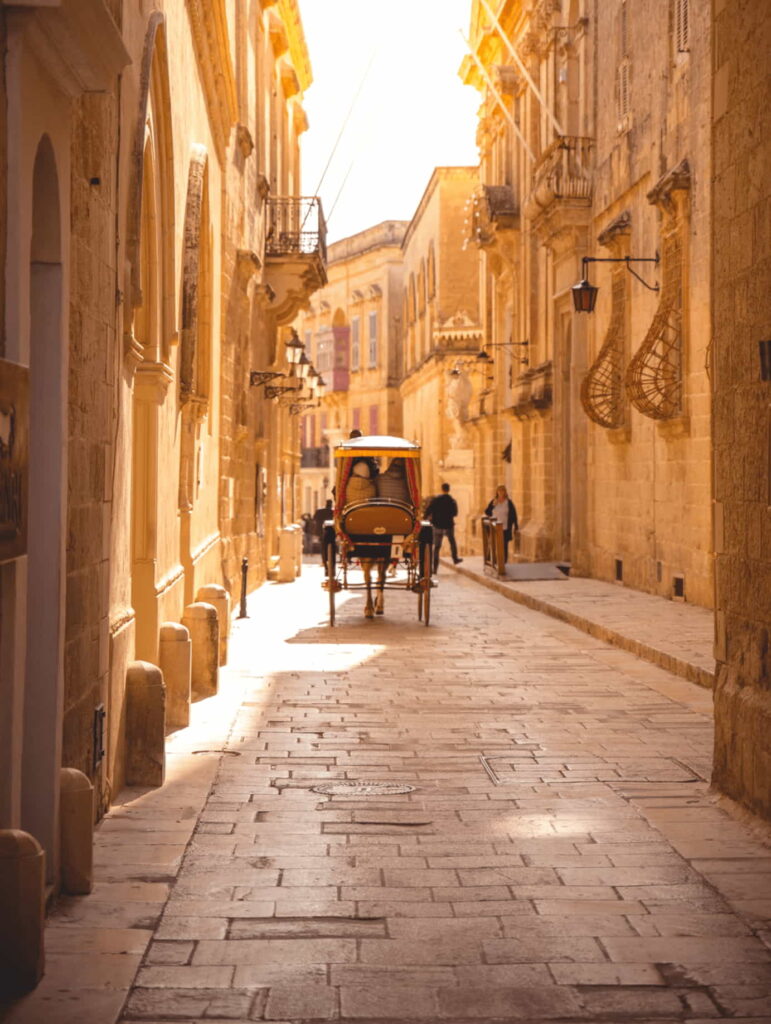 A horse-drawn karozzin traversing the cobblestone streets of Mdina, Malta, against a backdrop of medieval architecture and fortified walls. The golden sunlight illuminates the scene, casting long shadows as the traditional Maltese carriage adds a touch of nostalgia to the historic ambiance of the Silent City.