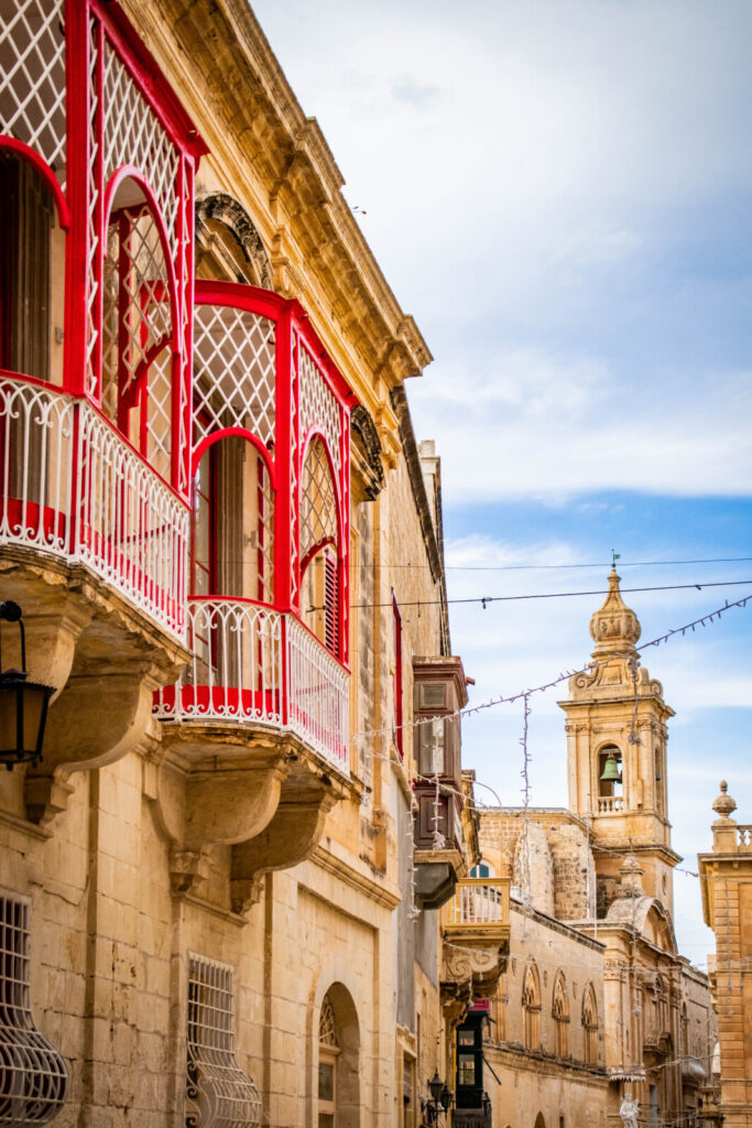 An atmospheric capture of an ancient building nestled within the fortified walls of Mdina, Malta. The weathered stone facade bears the marks of centuries, with ornate details and arched windows hinting at its rich history. Shadows play across the textured surface, evoking a sense of mystery and timelessness in the heart of the Silent City.