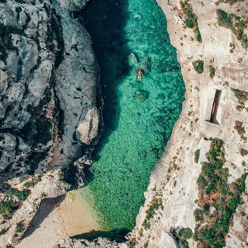 Wied il-Għasri, Gozo: A picturesque coastal chasm with narrow turquoise waters leading to a sandy beach, framed by rocky cliffs and lush greenery.