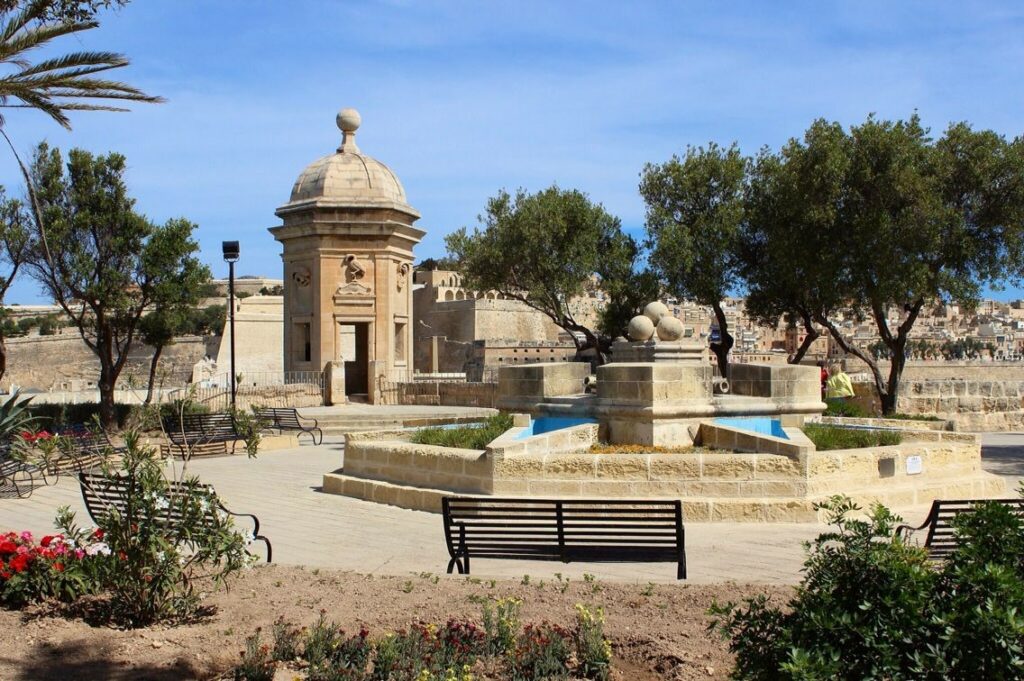 Gardjola Gardens, a serene oasis nestled within the fortified walls of Senglea, Malta. Lush greenery and vibrant flowers adorn the well-manicured garden, providing a peaceful retreat from the bustling city streets. A stone pathway meanders through the garden, leading visitors past charming benches and shaded alcoves perfect for relaxation. At the center of the garden stands the iconic Gardjola Watchtower, its sturdy walls adorned with the symbolic "eye" and "ear" sculptures that have become synonymous with Senglea's maritime heritage. From this vantage point, visitors can enjoy sweeping views of the Grand Harbour and beyond, making Gardjola Gardens a must-visit destination for nature lovers and history enthusiasts alike.