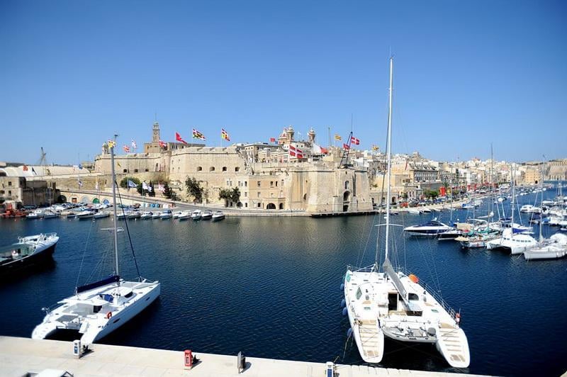 A sleek luxury yacht moored at the waterfront of Isla, Malta. The pristine white hull of the yacht gleams in the sunlight, reflecting off the crystal-clear waters of the Mediterranean Sea. In the background, the historic buildings of Isla create a striking backdrop, juxtaposing modern luxury with the city's rich maritime history.