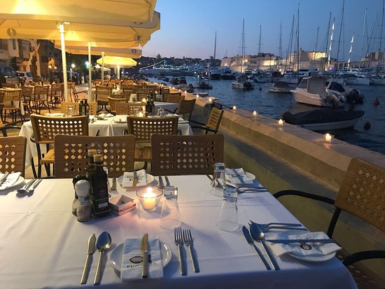 A picturesque outdoor dining area at Enchante Restaurant in Senglea, Malta. The restaurant's terrace overlooks the historic Senglea Marina, offering stunning views of the harbor and surrounding cityscape. Elegant tables and chairs are arranged neatly on the terrace, adorned with crisp white tablecloths and sparkling glassware. Colorful flower arrangements add a touch of natural beauty to the scene, creating a romantic and inviting ambiance for diners to enjoy their meals al fresco.





