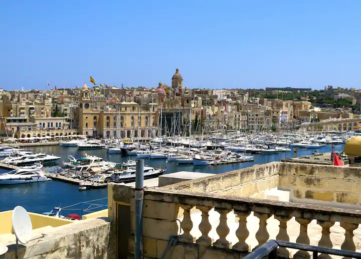 A picturesque view of Isla Marina in Senglea, Malta. Colorful fishing boats and sleek yachts bob gently in the shimmering waters of the Grand Harbour, their reflections dancing in the sunlight. Quaint waterfront cafes and restaurants line the promenade, their outdoor seating areas bustling with activity. Historic buildings and fortifications overlook the marina, adding a sense of timeless charm to the scene. Against the backdrop of the azure Mediterranean Sea, Isla Marina invites visitors to immerse themselves in the vibrant maritime atmosphere of Senglea and experience the beauty of Malta's coastal heritage.