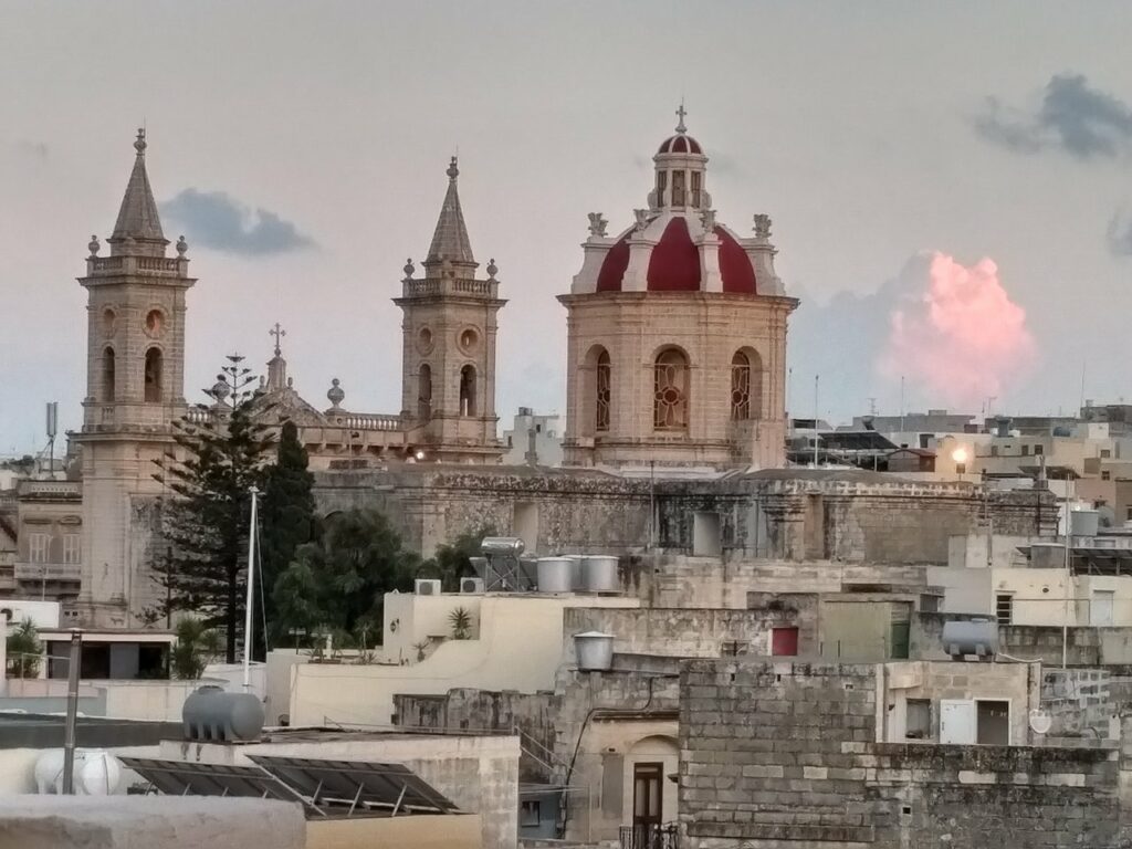 An aerial view of Qormi's skyline, showcasing the town's historic architecture and bustling streets intertwined with lush greenery, under a clear blue sky. The iconic domes of Qormi's churches stand tall amidst the surrounding buildings, providing a glimpse into the town's rich cultural heritage.