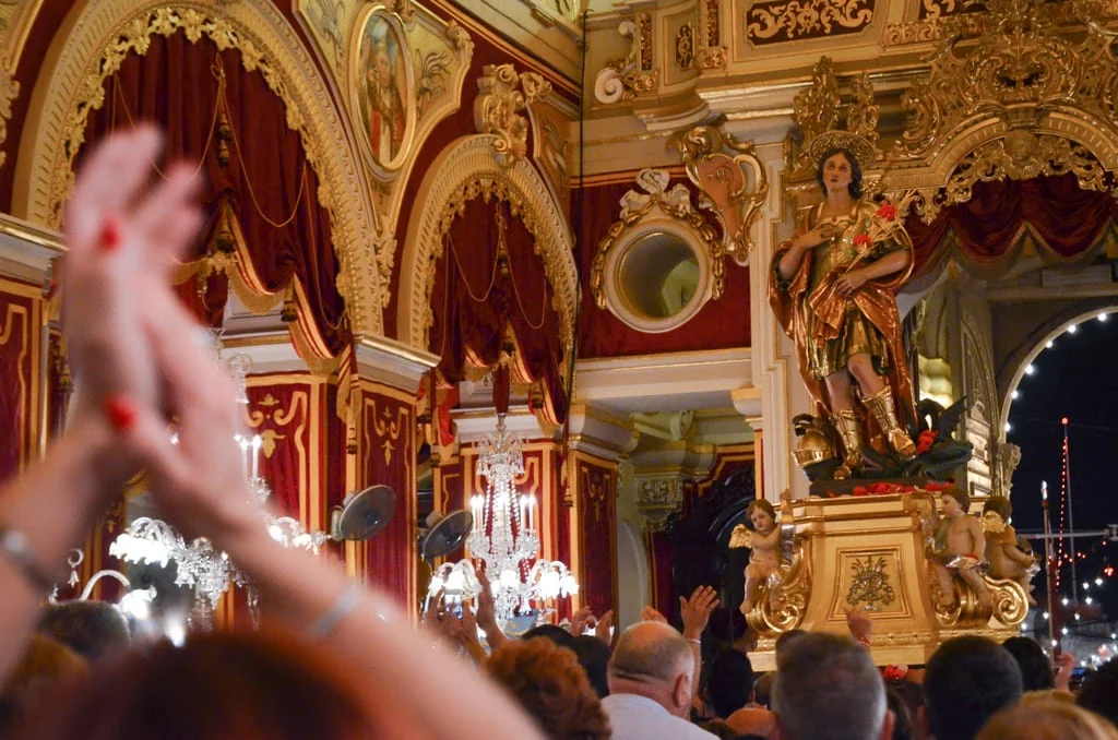 A stunning display of intricate decorations adorning the interior of a church in Qormi, Malta. Delicate floral arrangements, ornate candlesticks, and vibrant tapestries embellish the sacred space, creating a reverent atmosphere of beauty and devotion. The interplay of light and shadow accentuates the exquisite details, enhancing the spiritual experience for worshippers and visitors alike.