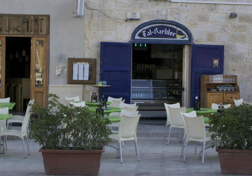 The charming entrance of Tal-Barklor restaurant in Senglea, Malta. A wooden door adorned with traditional Maltese wrought ironwork welcomes guests into the cozy eatery. Lush greenery surrounds the entrance, adding a touch of natural beauty to the facade. The warm glow of lights illuminates the doorway, inviting diners to step inside and experience the authentic Maltese hospitality and delicious cuisine that awaits within.