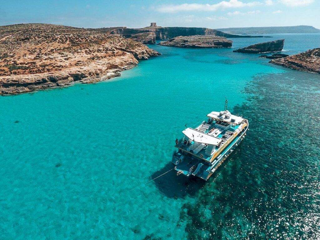 Comino Blue Lagoon Aerial View: A stunning aerial view of Comino's Blue Lagoon, showcasing its crystal-clear turquoise waters and white sandy seabed, perfect for swimming and snorkelling.